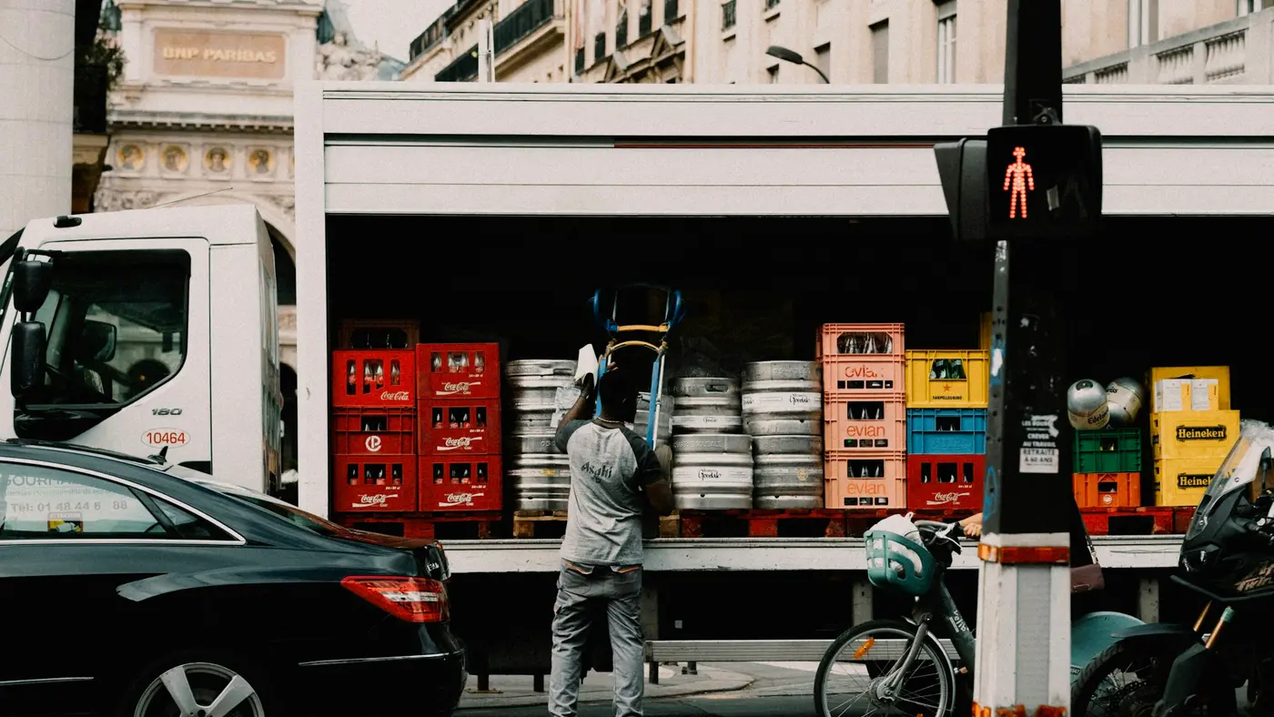 Entregador de distribuidora descarregando engradados de bebida do caminhão com o carrinho, numa rua da cidade