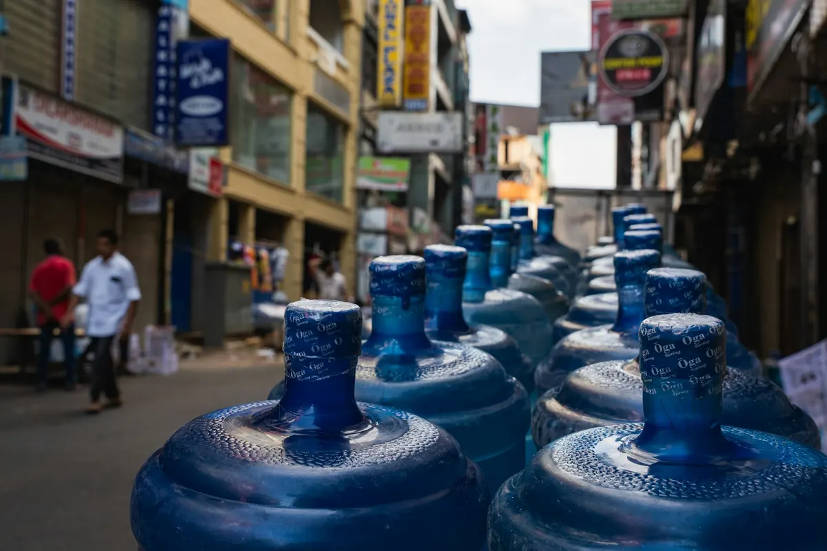 Galões de água de 20 litros enfileirados na calçada de uma rua comercial, prontos para a entrega